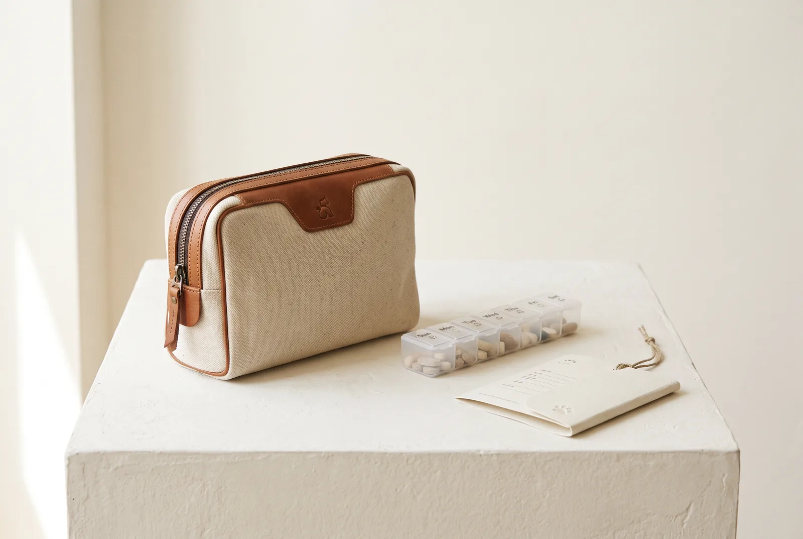 Canvas travel pouch, medication organizer, and emergency card on an ivory plinth